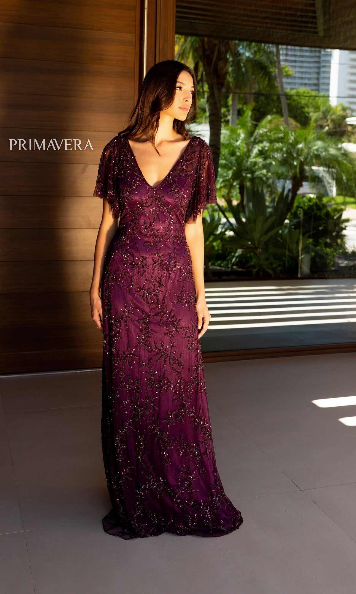 A woman stands indoors wearing the Cape-Sleeve Beaded Formal Dress: Primavera 13124, a floor-length deep purple gown with beaded details and cape sleeves. Sunlight from a window highlights the elegance of this formal dress.